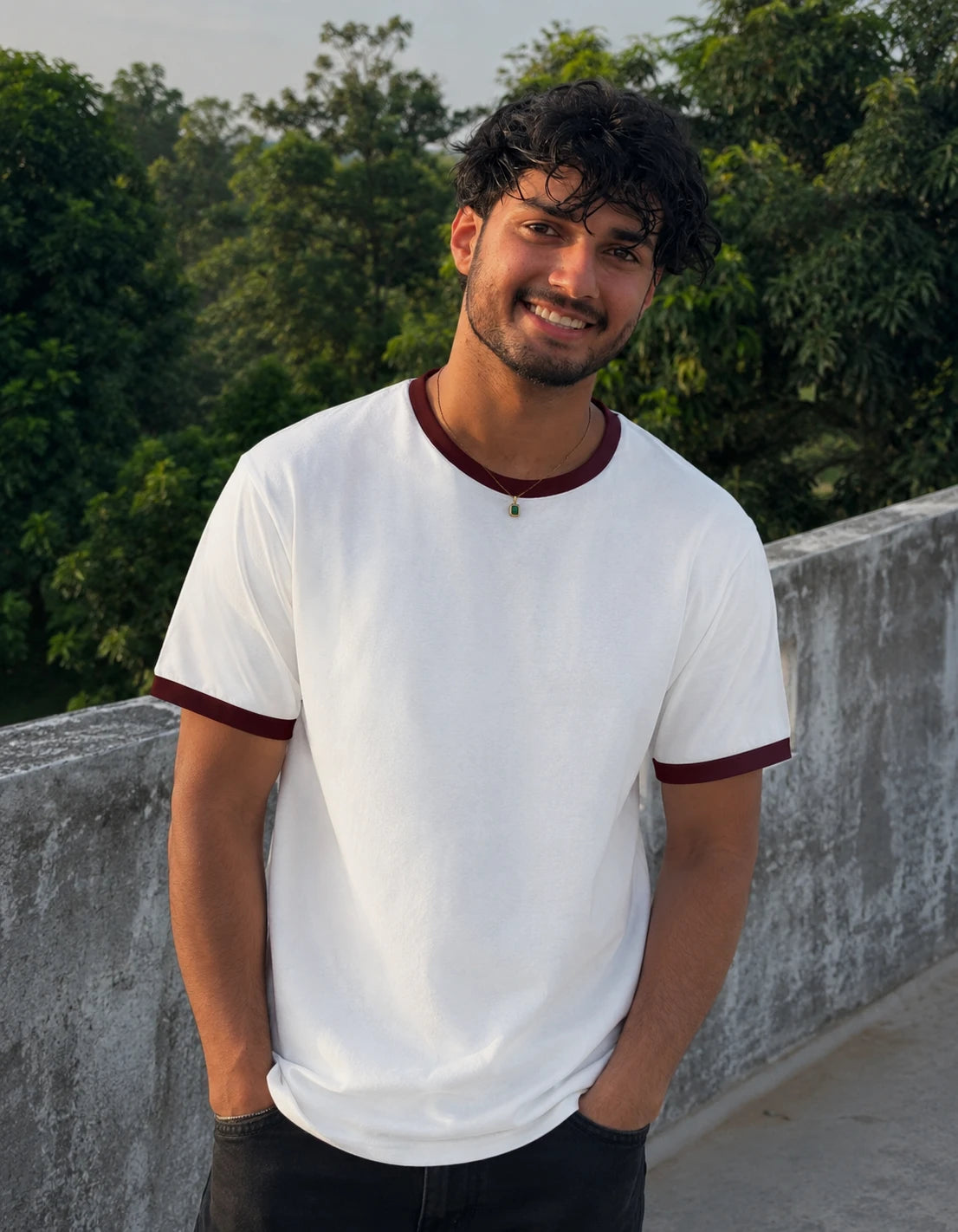 Man wearing a white t-shirt with maroon trim standing outdoors with greenery in the background