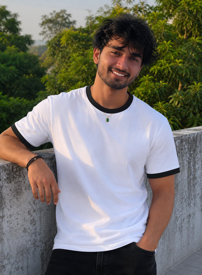 Man wearing a white t-shirt with black trim, leaning against a concrete wall with greenery in the background.