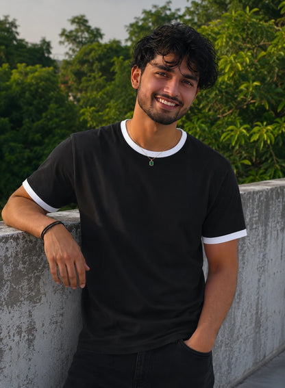 Man wearing a black t-shirt with white trim, standing outdoors with greenery in the background