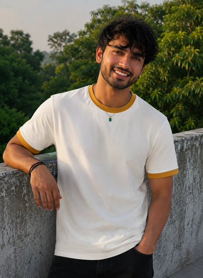 Man wearing a white t-shirt with yellow accents leaning against a wall with greenery in the background